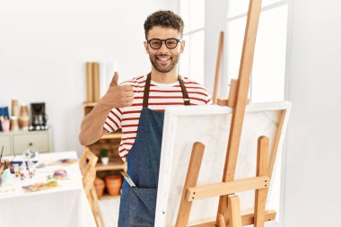 Sanat stüdyosundaki Arap genç adam elleriyle mutlu bir jest yapıyor. Kameraya bakıp başarıyı gösteren ifade. 