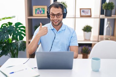 Hispanic man working using computer laptop wearing headphones smiling happy and positive, thumb up doing excellent and approval sign 