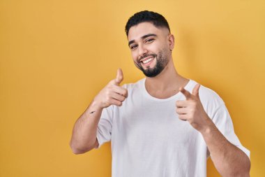 Sarı arka plan üzerine gündelik tişört giyen genç yakışıklı adam mutlu ve komik yüzlü parmaklarını kameraya doğrultuyor. iyi enerji ve titreşimler. 