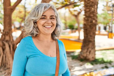 Orta yaşlı, kır saçlı, gülümseyen bir kadın şehirde mutlu mesut duruyor..