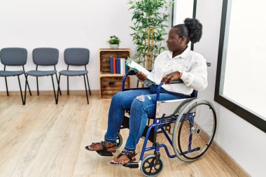 Bekleme odasında tekerlekli sandalyede oturan genç siyahi kadın yan yan yan bakıyor, rahat bir poz veriyor ve kendine güvenen bir gülümseme takınıyor.. 