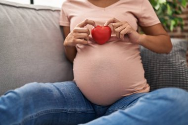 Kalbini taşıyan hamile genç Latin kadın evdeki kanepede oturuyor.