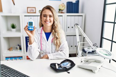 Young beautiful doctor woman holding glucose meter looking positive and happy standing and smiling with a confident smile showing teeth 