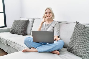 Young caucasian woman using laptop at home sitting on the sofa doing happy thumbs up gesture with hand. approving expression looking at the camera showing success. 
