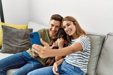 Young hispanic couple make selfie by the smartphone sitting on the sofa with dog at home.