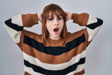 İzole edilmiş arka planda çizgili süveter giyen genç ve güzel bir kadın çılgın ve elleri başında korkmuş, korkmuş ve ağzı açık şoktan şaşırmış. 