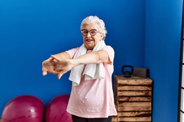 Spor kıyafetleri giyen yaşlı gri saçlı kadın spor merkezinde kendine gülerek duruyor.