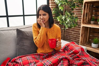 Kanepede oturan genç İspanyol kadın evde kahve içiyor. Gergin ve gergin görünüyor. Elleri ağızdan tırnak yiyor. anksiyete. 