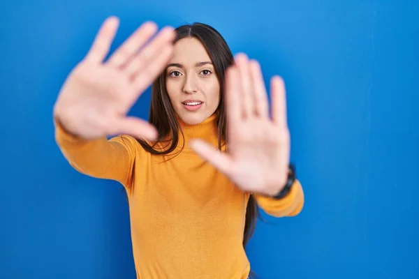 Young brunette woman standing over blue background doing frame using hands palms and fingers, camera perspective