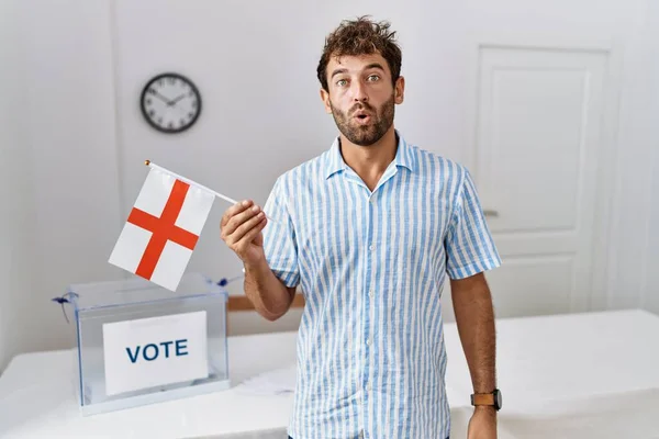 Young handsome man at political campaign election holding england flag scared and amazed with open mouth for surprise, disbelief face