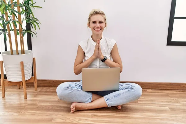 Young blonde woman using computer laptop sitting on the floor at the living room praying with hands together asking for forgiveness smiling confident.