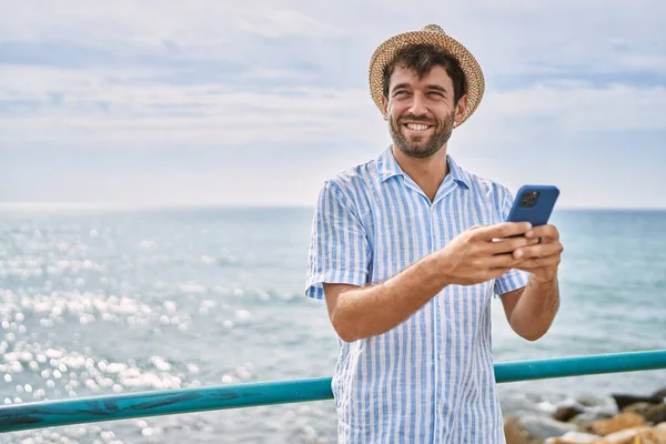 Genç İspanyol adam sahilde akıllı telefon kullanarak mutlu bir şekilde gülümsüyor.