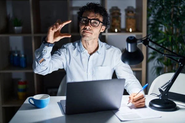 Hispanic man working at the office at night smiling and confident gesturing with hand doing small size sign with fingers looking and the camera. measure concept.