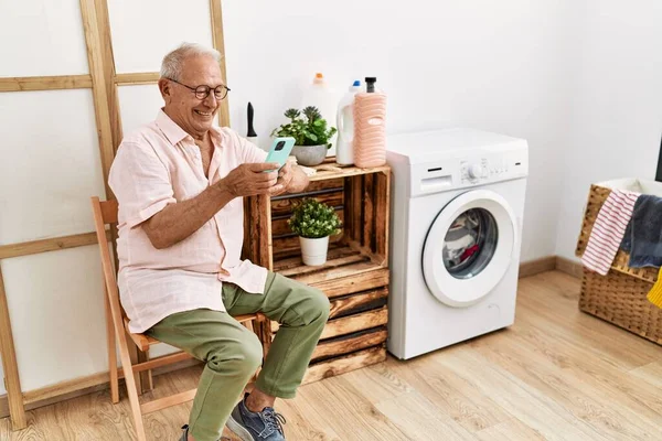 Senior man using smartphone waiting for washing machine at laundry room