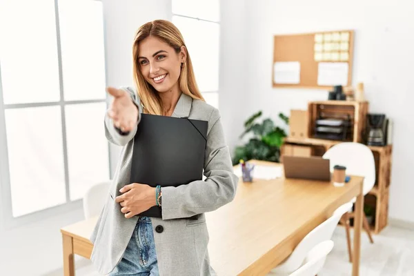 Blonde business woman at the office smiling cheerful offering palm hand giving assistance and acceptance.