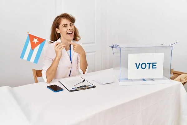 Beautiful caucasian woman at political campaign election holding cuba flag pointing thumb up to the side smiling happy with open mouth