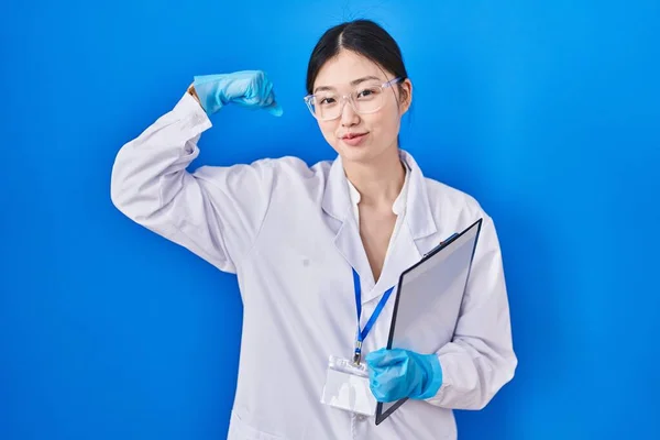 Chinese young woman working at scientist laboratory strong person showing arm muscle, confident and proud of power