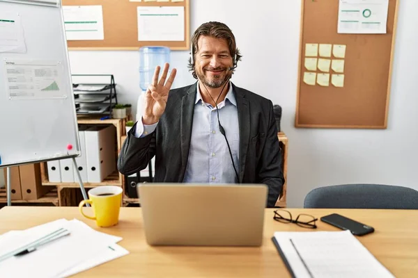 Handsome middle age man wearing call center agent headset at the office showing and pointing up with fingers number three while smiling confident and happy.