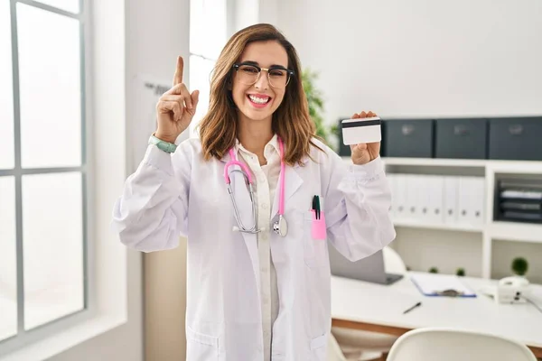 Young doctor woman holding credit card at the clinic smiling with an idea or question pointing finger with happy face, number one