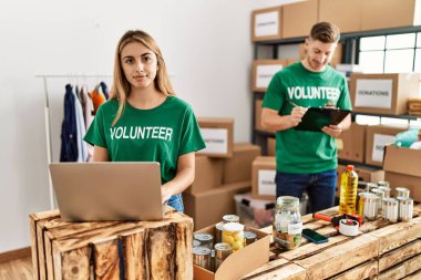 Young woman and man wearing volunteer t shirt at donations stand thinking attitude and sober expression looking self confident 