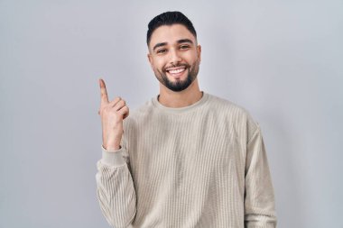 Genç ve yakışıklı bir adam izole edilmiş arka planda neşeli, yüzünde bir gülümsemeyle, mutlu ve doğal bir ifadeyle elini ve parmağını kaldırmış duruyor. 