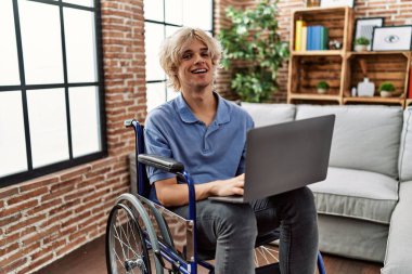 Genç sarışın adam evde tekerlekli sandalyede dizüstü bilgisayar kullanıyor.