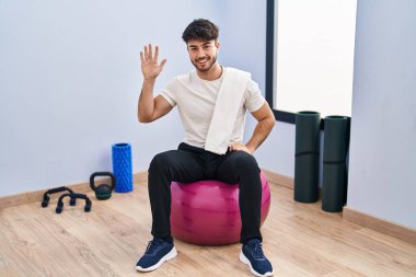 Hispanic man with beard sitting on pilate balls at yoga room showing and pointing up with fingers number five while smiling confident and happy. 