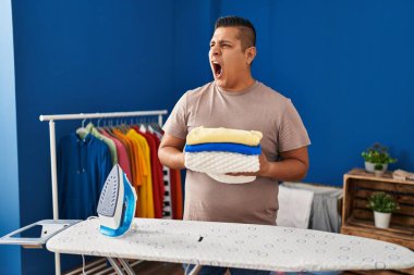 Hispanic young man holding folded laundry after ironing angry and mad screaming frustrated and furious, shouting with anger. rage and aggressive concept. 