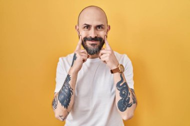 Young hispanic man with tattoos standing over yellow background smiling with open mouth, fingers pointing and forcing cheerful smile 