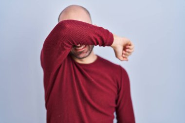 Beyaz arka planda duran sakallı genç kel bir adam gözlük takıyor kollarıyla gözleri örtüyor neşeli ve komik gülümsüyor. kör kavram. 