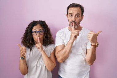 Middle age hispanic couple together over pink background asking to be quiet with finger on lips pointing with hand to the side. silence and secret concept. 