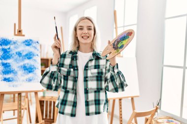 Resim stüdyosunda fırça ve palet tutarak mutlu gülümseyen genç sanatçı kız..