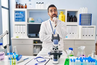 Laboratuvarda sakallı, eli çenesinde çalışan ve soru sormayı düşünen genç bir İspanyol. Düşünceli bir ifade. Düşünceli bir yüzle gülümsüyor. şüphe kavramı. 