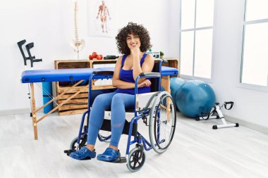 Young middle eastern woman sitting on wheelchair at physiotherapy clinic looking confident at the camera smiling with crossed arms and hand raised on chin. thinking positive. 