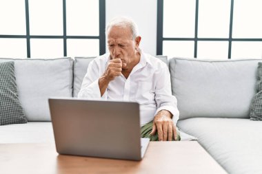 Senior man using laptop at home sitting on the sofa feeling unwell and coughing as symptom for cold or bronchitis. health care concept. 