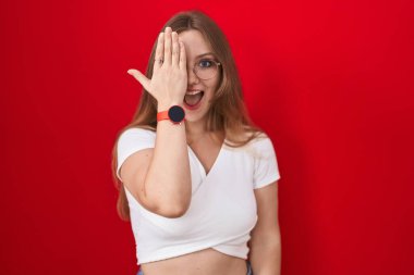 Genç beyaz kadın kırmızı arka planda duruyor, bir gözünü eliyle kapatıyor, yüzünde kendine güvenen bir gülümseme ve sürpriz bir duygu var.. 
