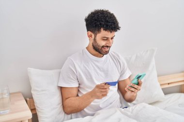 Genç Arap adam yatak odasında yatağında oturan akıllı telefon ve kredi kartı kullanıyor.