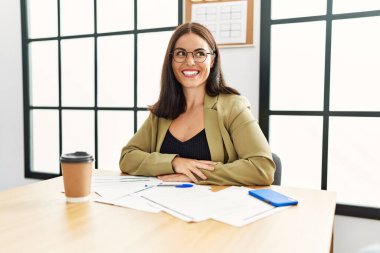 İş stili giyinmiş genç esmer kadın ofiste oturmuş yüzünde gülümseme ve doğal bir ifadeyle yan yana bakıyor. Kendinden emin gülen. 