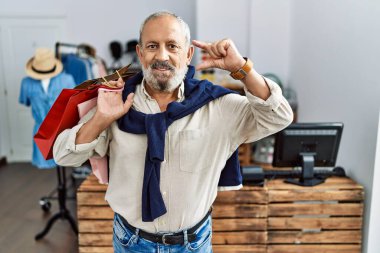 Handsome senior man holding shopping bags at boutique shop smiling and confident gesturing with hand doing small size sign with fingers looking and the camera. measure concept. 