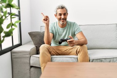 Orta yaşlı İspanyol bir adam video oyunu oynuyor kanepeye oturmuş gülümsüyor mutlu bir şekilde elini ve parmağını kenarı gösteriyor. 