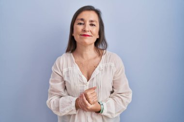 Middle age hispanic woman standing over blue background with hands together and crossed fingers smiling relaxed and cheerful. success and optimistic 