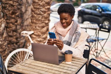 Laptop ve akıllı telefon kullanan Afro-Amerikalı bir kadın kahve dükkanının terasında oturuyor.