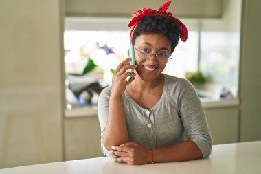 Afro-Amerikalı kadın akıllı telefonla konuşuyor. Evde masada oturuyor.