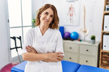 Orta yaşlı fizyoterapist kadın ağrı kesici kliniğinde çalışıyor. Kollarını kavuşturup kameraya bakarken gülümsüyor. pozitif kişi. 