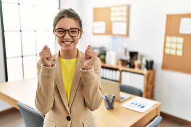 Young brunette teenager wearing business style at office excited for success with arms raised and eyes closed celebrating victory smiling. winner concept. 