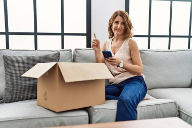 Orta yaşlı bir kadın koltukta oturmuş telefondaki teslimat paketini kontrol ediyor. Bir fikir ya da soru işareti ile şaşırmış bir şekilde mutlu bir yüzü işaret ediyor. 