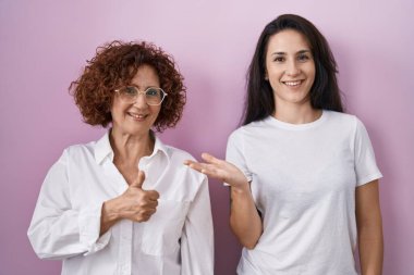 İspanyol anne ve kızı pembe arka plan üzerine gündelik beyaz tişört giyip avuç içini gösteriyor ve baş parmaklarıyla iyi bir jest yapıyor, mutlu ve neşeli gülümsüyor. 