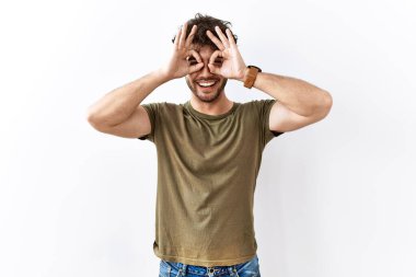 Hispanic man standing over isolated white background doing ok gesture like binoculars sticking tongue out, eyes looking through fingers. crazy expression. 