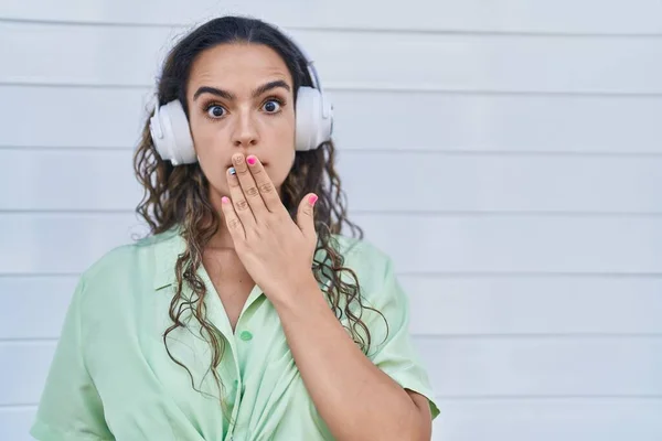 若いヒスパニック系の女性は 手で口をカバーヘッドフォンを使用して音楽を聞いてショックを受け 間違いを恐れている 驚きの表情 — ストック写真