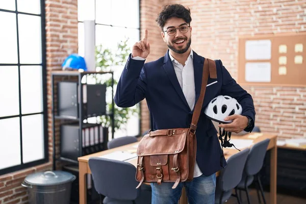 Hispanic man with beard holding bike helmet at the office surprised with an idea or question pointing finger with happy face, number one
